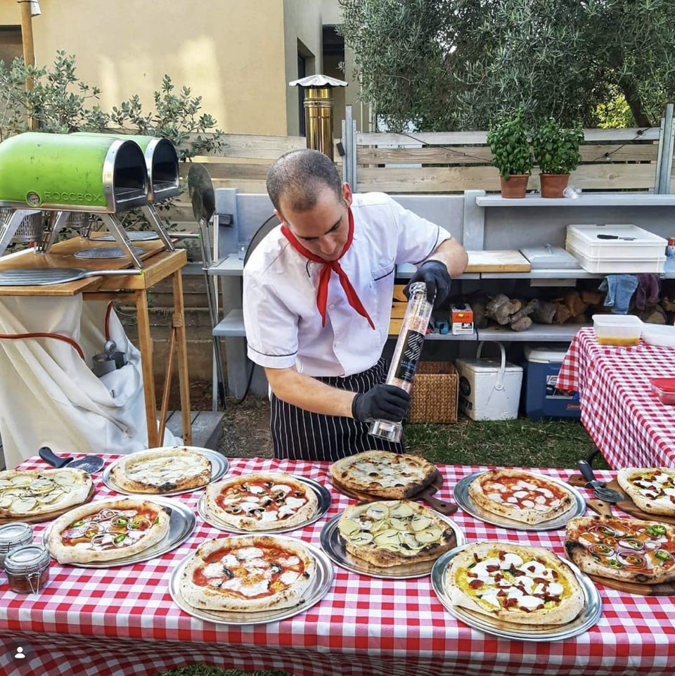 Alon seasoning pizzas at an outdoor event, two portable ovens behind him